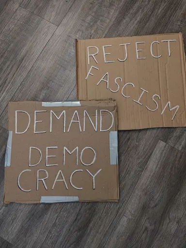 Two simple cardboard signs with black and white lettering. One sign reads "DEMAND DEMOCRACY" and the other "REJECT FASCISM" in all capital letters