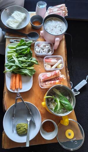 A top down photo of a yellow and pink hotpot on a wooden coffee table, with various noodles, vegetables such as bok choy and carrot, lamb and beef rolls, prawns. There is an electrical powerboard sitting in front of the table, right underneath the spill zone.